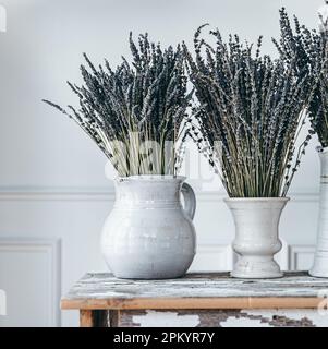 White ceramic vases with branches of blooming lavender flowers placed on wooden table against light wall Stock Photo