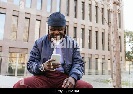 Positive black male with dreadlocks sitting and browsing smartphone while resting on city street in daylight Stock Photo