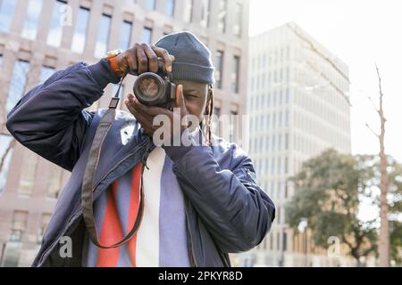 Black male photographer with professional photo camera standing against building and taking picture in city street Stock Photo