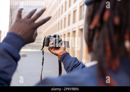 Back view black young male in trendy clothes with braids standing outside and looking at camera while taking self photo on professional camera Stock Photo