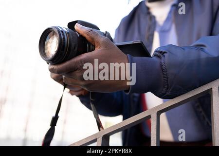 Crop black male photographer with professional photo camera standing against building and taking picture in city street Stock Photo