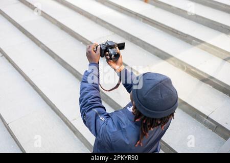 From above black male in trendy clothes with braids standing outside and looking at camera while taking self photo on professional camera Stock Photo