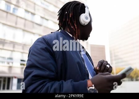 Young black male with earphones looking away while standing and listening to music on city street Stock Photo