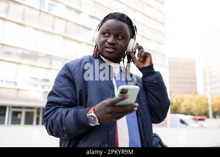Young black male with earphones looking away while standing and listening to music on city street Stock Photo