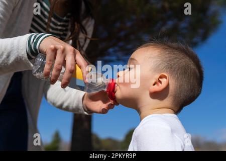 Side view of little boy drinking fresh water from plastic bottle while crop mother helping during walk outside Stock Photo