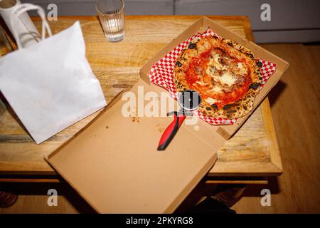 From above of appetizing baked pizza with tomatoes and cheese with crispy crust placed in cardboard box with knife on table Stock Photo