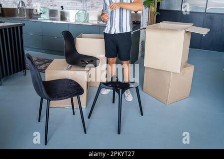 Anonymous male in casual clothes standing near cardboard boxes and assembling chair while moving into new apartment Stock Photo