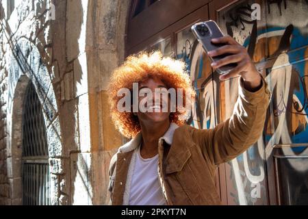 Positive young ethnic male with Afro hairstyle taking self portrait while looking at screen of mobile phone and standing near wall with graffiti in su Stock Photo