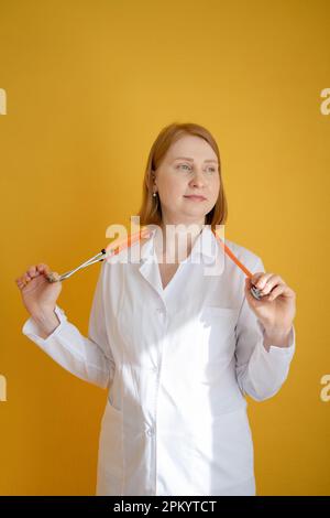 Smiling young female doctor in white coat using stethoscope and looking away while standing against yellow background Stock Photo