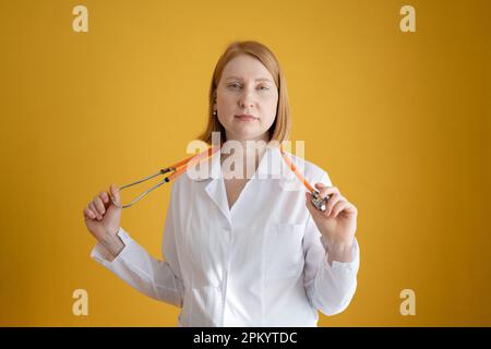 Smiling young female doctor in white coat using stethoscope and looking at camera while standing against yellow background Stock Photo