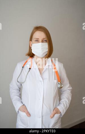 Young female doctor standing in medical coat and protective mask and stethoscope behind neck with both hands in pocket and looking at camera against g Stock Photo