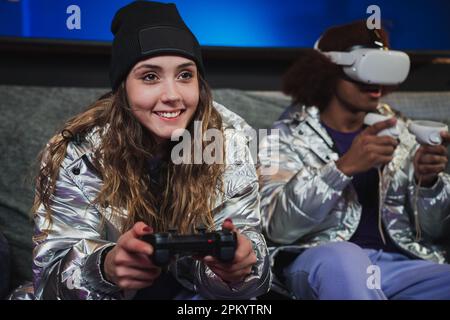 Young woman and anonymous friend in VR glasses sitting on couch together and playing video game with joysticks at home Stock Photo