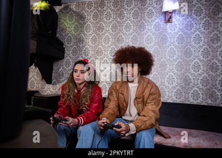 Concentrated curly Hispanic guy and female partner sitting on couch with controllers and entertaining with video game in living room Stock Photo