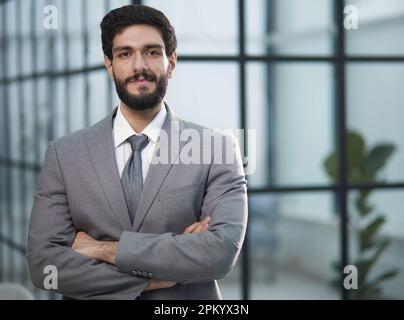 a dark-haired man in an office in a suit stands with crossed arms in front of him Stock Photo