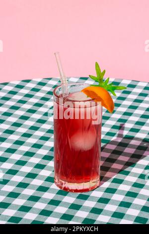 Glass of cold refreshing red cocktail with ice and straw served with orange on table with checkered tablecloth Stock Photo