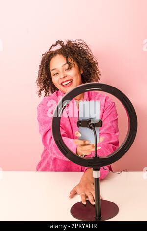 Cheerful young ethnic curly haired female blogger in pink shirt recording video on smartphone while smiling and using ring lamp Stock Photo