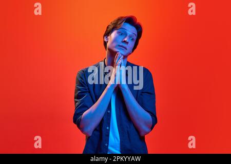 Studio shot of young guy, student looking with compassionate eyes over red-orange background in neon light. Concept of beauty, youth, human emotions Stock Photo