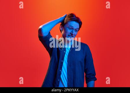 Portrait of young guy, man wearing shirt standing with uncertain facial expression over red-orange background in neon light. Doubtful guy Stock Photo