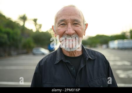 Portrait of smiling senior bearded male in casual clothes looking at camera while standing on blurred city street during daytime Stock Photo