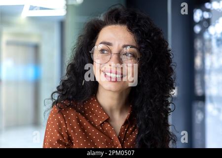 Close-up photo. Portrait of young hispanic woman with glasses and curly hair sitting, working and studying in office, campus. Smiling, she looks to the side. Stock Photo