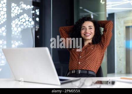 A young beautiful Latin American woman sitting in the office on a chair and resting, taking a break, satisfied with the work done. She threw her hands behind her head, smilingly looks to the side. Stock Photo