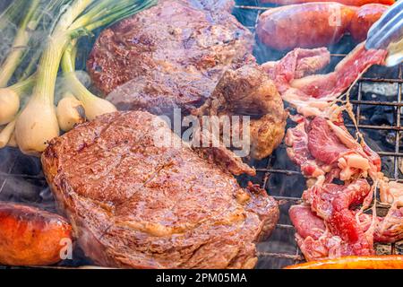 Delicious Mexican barbecue with beef steaks, chorizo and white onions grilled over charcoal in a brazier, smoke coming out, thin and thick steaks, met Stock Photo