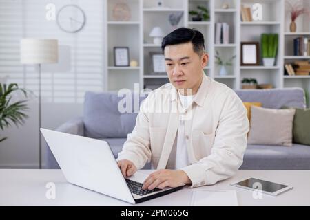 Serious and focused Asian man, businessman, freelancer, student working studying at home using laptop, Sitting at table and talking on video call, typing. Stock Photo