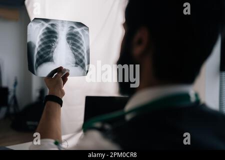 Closeup back view of unrecognizable male doctor examining patient chest x-ray film lungs scan at radiology department in hospital sitting at desk with Stock Photo