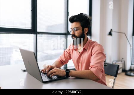 Focused Indian businessman wearing casual clothes working typing on laptop sitting at table in light office room on background of window. Stock Photo