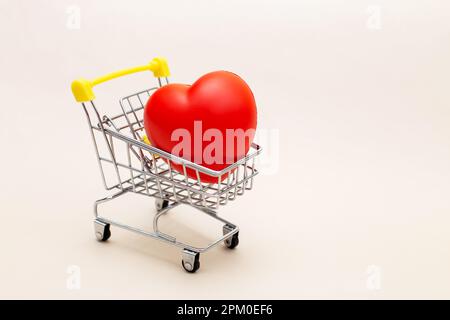 Red heart in the shopping cart or trolley isolated on beige background. Shopping for wedding, womens day concept, gifts for Valentines Day. Stock Photo