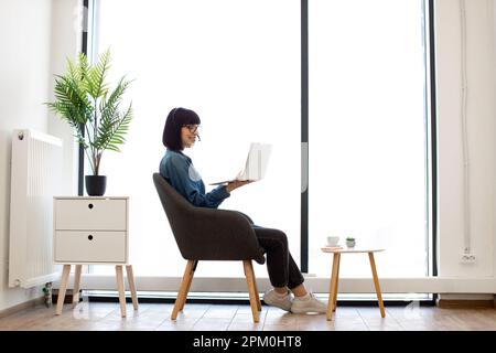 Pretty young female in spectacles handling modern laptop while sitting near coffee table in creative workplace. Cheerful businesswoman in casual outfit holding video conference via wireless headset. Stock Photo