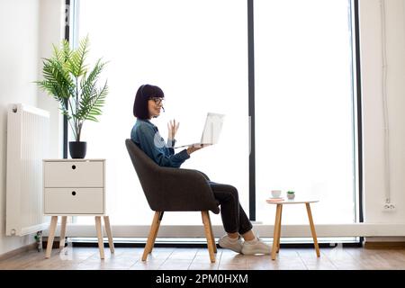 Pretty young female in spectacles handling modern laptop while sitting near coffee table in creative workplace. Cheerful businesswoman in casual outfit holding video conference via wireless headset. Stock Photo