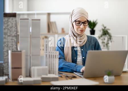 Focused bespectacled muslim female in hijab typing on portable computer at office desk with building models on it. Experienced architect developing urban design using modern gadgets and drawing tools. Stock Photo