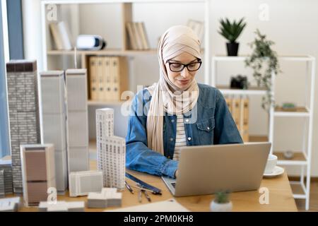 Focused bespectacled muslim female in hijab typing on portable computer at office desk with building models on it. Experienced architect developing urban design using modern gadgets and drawing tools. Stock Photo