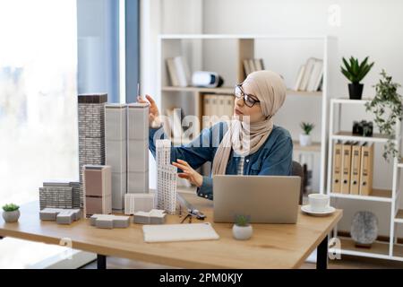 Beautiful arabian female in hijab and glasses arranging building models on office desk during coffee break in creative workplace. Architectural designer analysing details of new project using laptop. Stock Photo