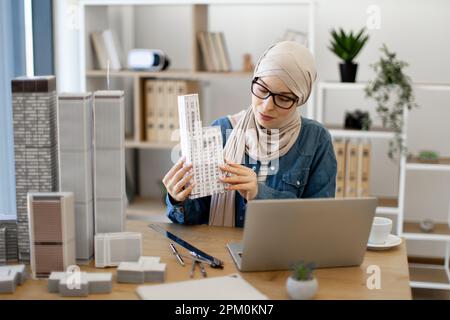 Thoughtful muslim lady in hijab directing building model at laptop camera while working on urban design project in modern workspace. Experienced engineer holding online briefing via video call app. Stock Photo