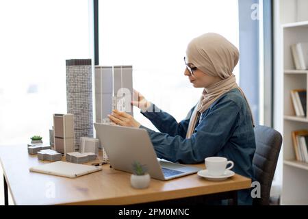 Thoughtful muslim lady in hijab placing architectural models on desk surface near smart gadgets in creative workplace. Residential architect analysing project data using wireless technologies. Stock Photo