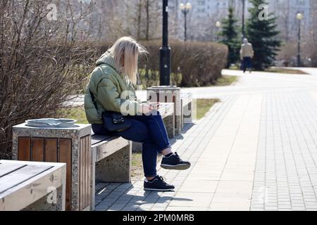 Blonde woman in jacket and jeans sitting on wooden bench and using smartphone, leisure in city park at spring Stock Photo