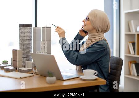 Cheerful arabian female wearing hijab returning phone call to colleague while using portable computer for innovative project in office. Talented designer finding inspiration with pencil in hand. Stock Photo