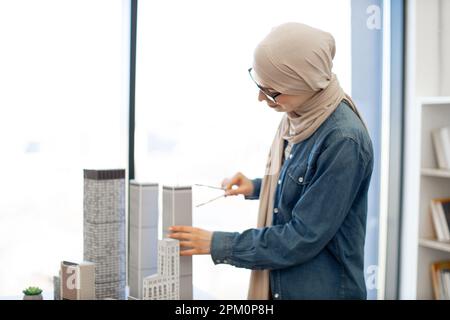 Arabian businesswoman in hijab and spectacles examining building model with compass while standing near writing desk with coffee and gadgets on it. Talanted architect using drawing tools at work. Stock Photo