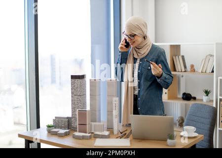 Muslim female worker in hijab and glasses having pleasant talk over smartphone while standing near office chair and keeping drawing pencil in hand. Smiling architect discussing construction project. Stock Photo