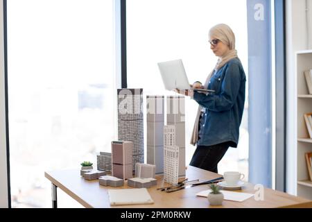Muslim female worker in hijab and casual clothes working on portable computer while standing near office desk with architectural models. Talented designer creating new project plan on laptop. Stock Photo