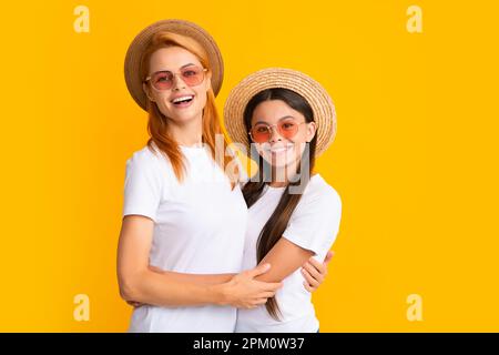 Happy stylish mother and daughter posing at studio yellow background, wearing straw hat and sunglasses. Summer family portrait. Stock Photo