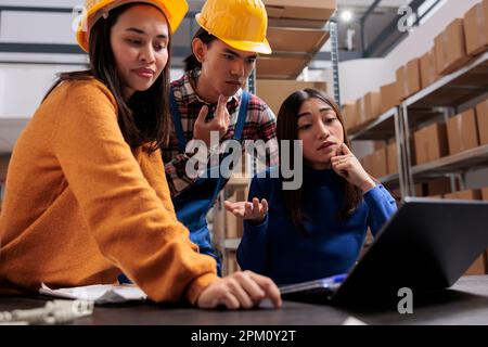 Asian warehouse managers team analyzing shipping checklist on laptop while standing near desk. Retail storehouse workers checking logistics plan on computer in storage room Stock Photo