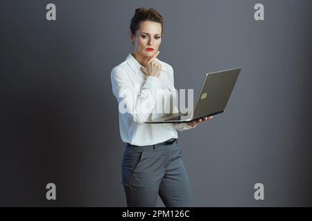 pensive young woman worker in white blouse using laptop isolated on grey. Stock Photo