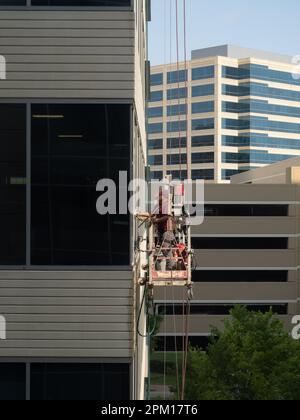 Eye level view of two male uniformed window washers cleaning glass on an office building while standing on a suspended scaffold. Stock Photo