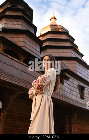 Beautiful woman wearing embroidered dress near old wooden church in village. Ukrainian national clothes Stock Photo