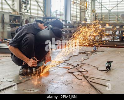 Male industrial worker wearing a weldig mask using an angle grinder on a steel plate, sparks flying. Stock Photo