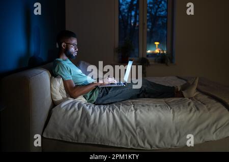 Black student guy lying in bed with laptop in evening, studying until late hours Stock Photo