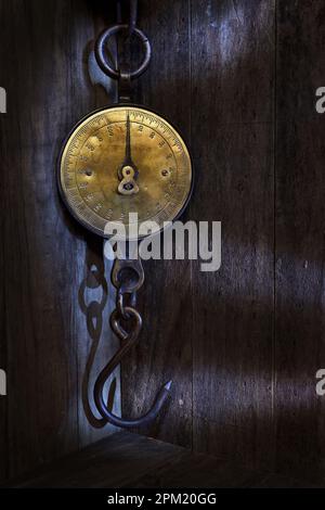 An antique spring balance, brass face, weighing scale with a hook, hanging in a dark, rustic setting in pools of mood lighting Stock Photo